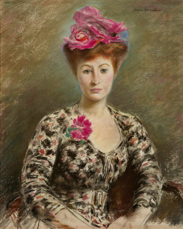 A woman sits with her hands in her lap. She is wearing a floral dress with a red flower at her neckline which match the red in her hat.