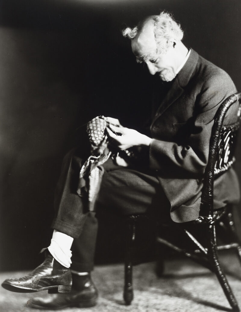 An elderly man sits in a wicker chair mending.