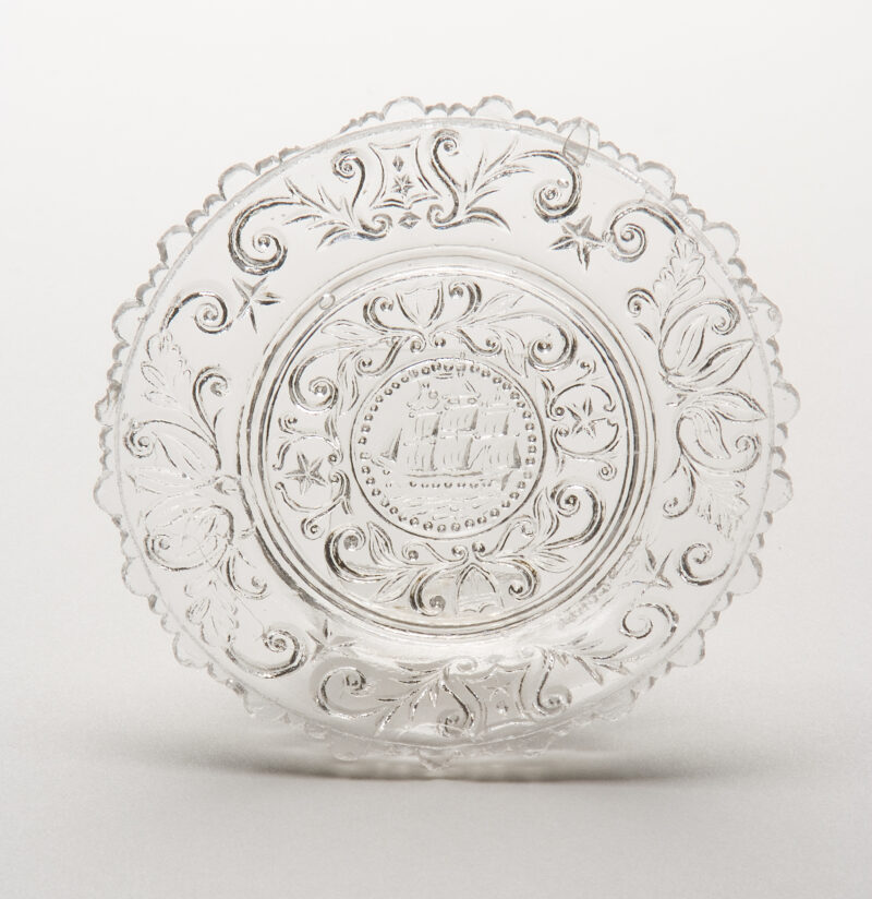 Circular plate with frigate at center encircled by a beaded ring and scroll design with shields & stars. Scroll design with shields & stars also around border. Rim has alternating pair of small scallops and single larger scallops.