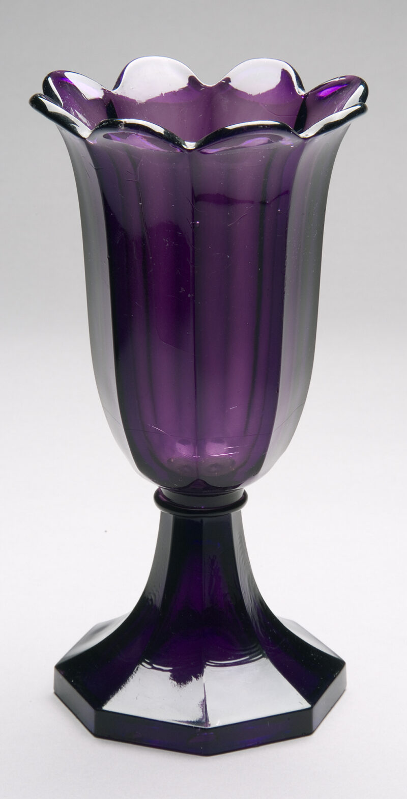 Trumpet-shaped vase with 8 vertical panels around sides. Rim scalloped & flared. Flat disk below bowl joins bowl to base. Flaring, hollow octagonal pedestal base. Transparent amethyst color.