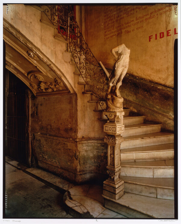 A stone stairway with a decorative railing with a headless figure on a pedestal at the first stair. The word Fidel is on the wall in red.