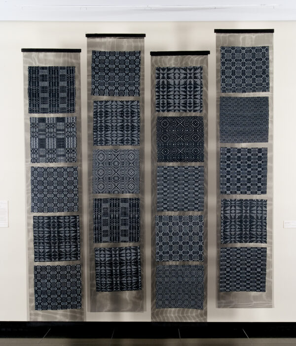 The object consists of 4 panels each having five squares of traditional weaving floated between two pieces of black window screen. Each square is an indigo and cream colored overshot weaving in a traditional American pattern. Each pattern is repeated twice in the total layout.