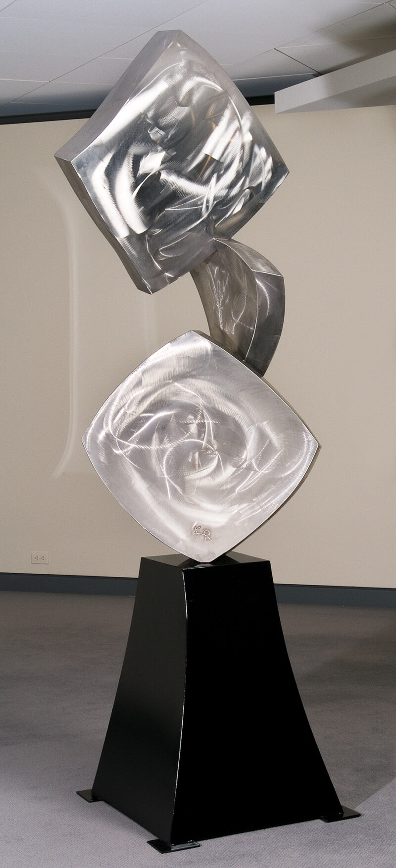 The sculpture is of three shapes: two squares with a triangle between. All of the shapes have a curve and have a random swirl surface treatment. These are vertical on a black base.