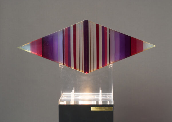 The sculpture is of poured liquid resin and then machined to its final form - the shape of a flattened triangle. It has vertical segments of color which changes from clear to pink to dark red and purple. The sculpture is supported on a clear plexi stand.