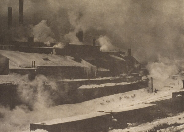 Scene of an industrial site with buildings and smoke stacks. There is snow on the ground and one figure on the road.
