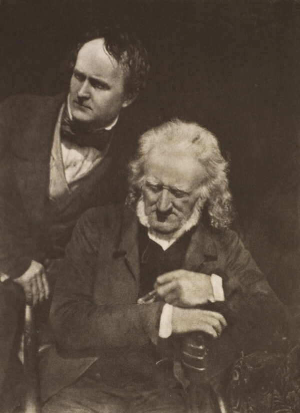 A portrait of two men. One standing looking to his right, the other is sitting. The seated man holds his glasses in his left hand and his right hand is just below his left resting on the arm of the chair.
