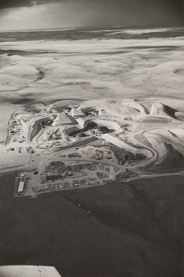 Round missile silos surrounded by banked earth and rolling empty hills in the background. The tip of the plane wing is visible at lower left.