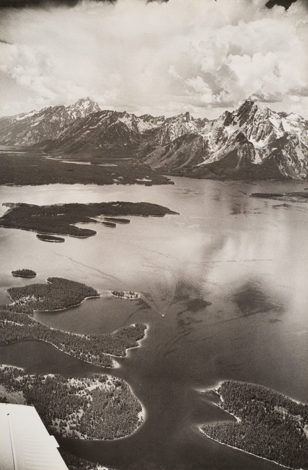 Lake with small islands and mountains with snow and cloud filled sky. Plane’s wing is visible at lower left.