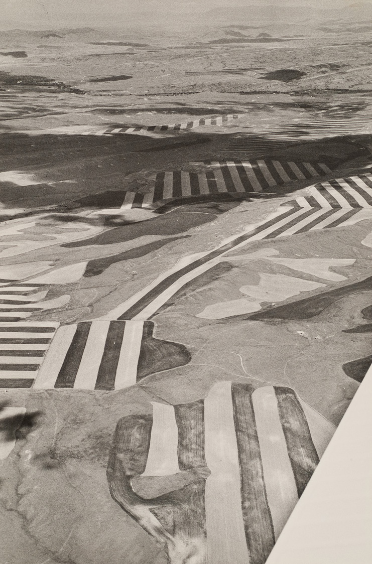 Patch work of fields with strong dark/light stripes. Wing of plane can be seen in lower right corner. Clouds are casting a shadow near the top.