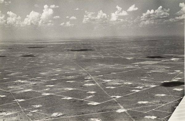 Flat land that is marked in a grid with white roads and white squares for oil production. Clouds are casting dark shadows on the land.