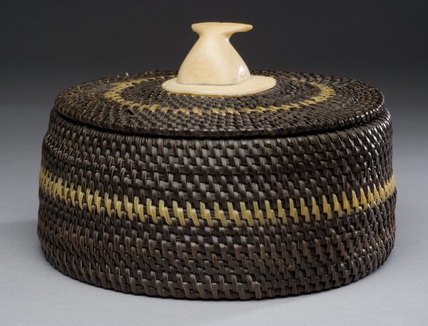 A lidded basket – both parts are dark brown each with a cream colored stripe. The basket has a 2 ј”  ivory disk at the center bottom and the lid has a 2 Ѕ” square with rounded corners. On top of this square is the tail of a whale as if diving underwater. The whale tail is pegged with wood to the lid.