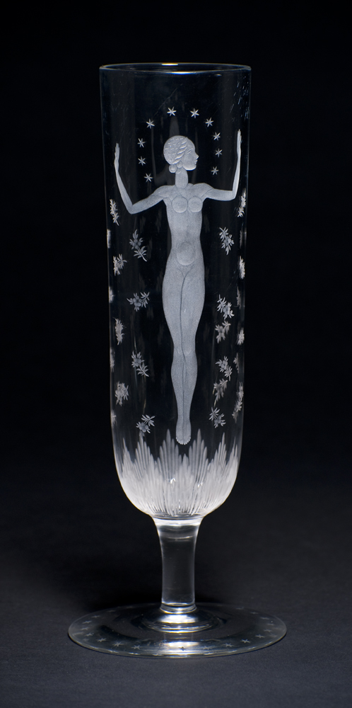 A vase with the image of a  female nude standing with arms up-raised. A floral motif surrounds her and near the bottom is stylized grass. The vase shape is a narrow column with a foot that is ringed with stars.