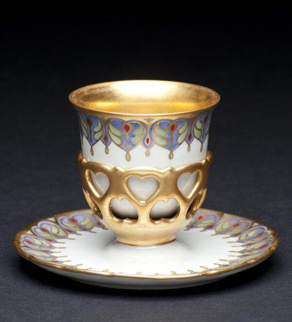 A cup without a handle, fits into a pierced gold design attached to a saucer. The saucer and cup have a decorative design in blue, green and red. The cup has a gold interior and the design is outlined in gold. The underside of the saucer is iridescent “rainbow” white.