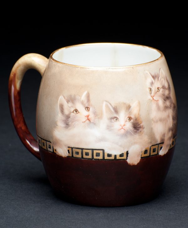 A mug decorated with three kittens on light brown background with dark brown on bottom half of mug separated by a row of gold squares.