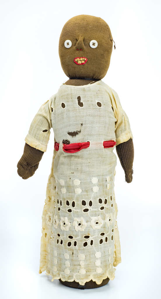 A freestanding female bottle doll. It has a glass bottle structure that is covered with stuffed black stockinette. The girl wears a white dress that is made of thin lace fabric with scalloped bottom edge. It is tied with a red silk ribbon tie at the waist. Button eyes and stitched facial features are present.