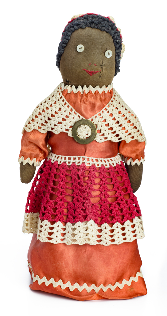 A female doll wears an orange dress with white rick rack trim, and red and white crocheted hat, shawl and apron. The eyes are buttons and the mouth and nose are embroidered. The hair is yarn. The doll has a bottle filled with sand for a stand, cotton stuffing. Her skin is fine brown stocking, with a larger texture stocking for the body. The under skitk is printed gingham with a crocheted trim around the bottom.