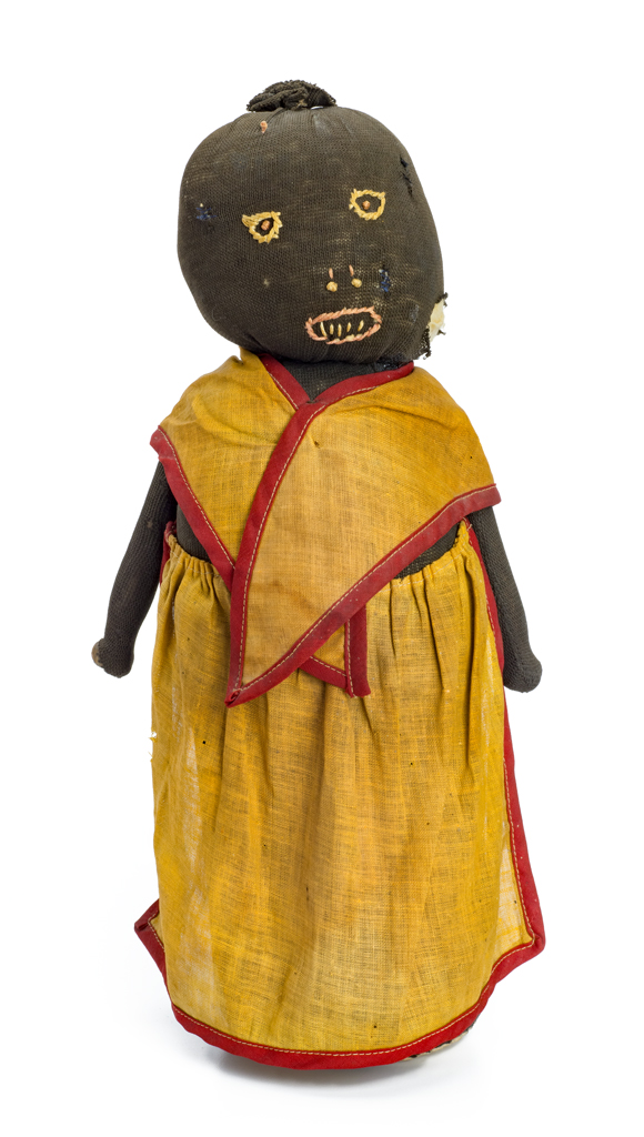 A female doll wears a tan skirt and shawl with red trim, over a bottle filled with sand. Her features are embroidered. The dark brown stocking is stuffed with cotton. She has wood dowels for hands.
