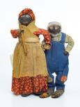 Possible hazel nut heads. The female wears a red print dress and head scarf with a brown apron and shoulder scarf. She carries a wood staff. The white-bearded man, wears blue overalls with patches at their knees. Both wear crepe paper shoes
