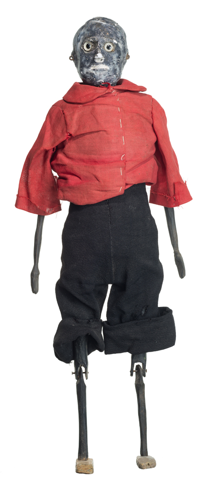 Puppet of male figure wears a red shirt and black pants. He has articulated arms and legs. His head is made of painted plaster with plastic moveable eyes and screws for ears