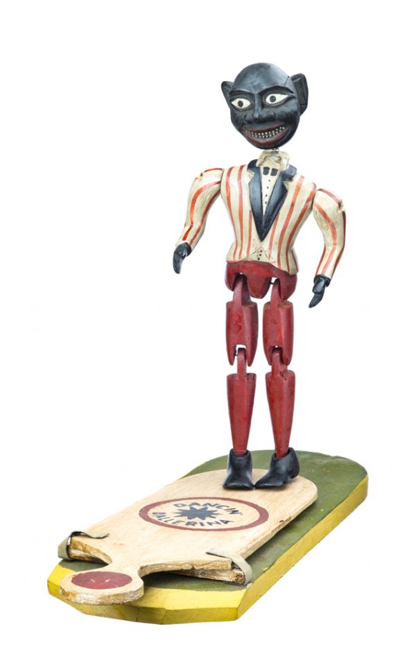 A wood doll with articulated joints and a spring head. He has red pants, a yellow and red stripped top coat with black collar.The wood and wire stand for the Dancing Ballerina is painted green, white and red. The base consists of a movable paddle atop the main platform that is secured with two leather straps. The user can press the end of the paddle to move the doll.