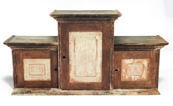 The object is a tiered cabinet with a flat base. It is made of painted wood and features a tall central compartment that is flanked by two shorter compartments. The surfaces are painted a rust color with white front panels.