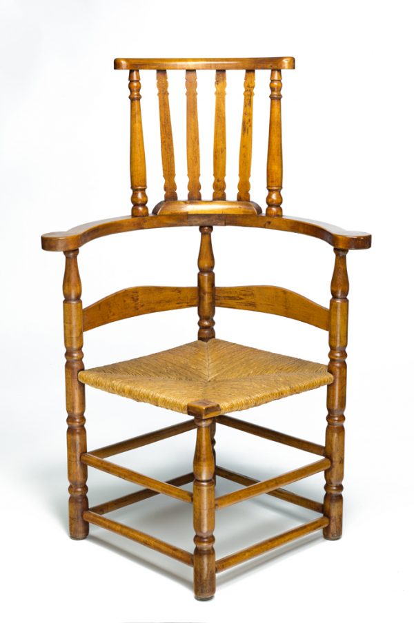 A corner chair with a curved pillow crest back and flat arms that terminate in an outward curl. A smaller banister-back section continues upward. It has turned legs and a woven fiber (rush) seat. Two stretchers connect each leg, and the seat is angled to allow the sitter to face either direction.
