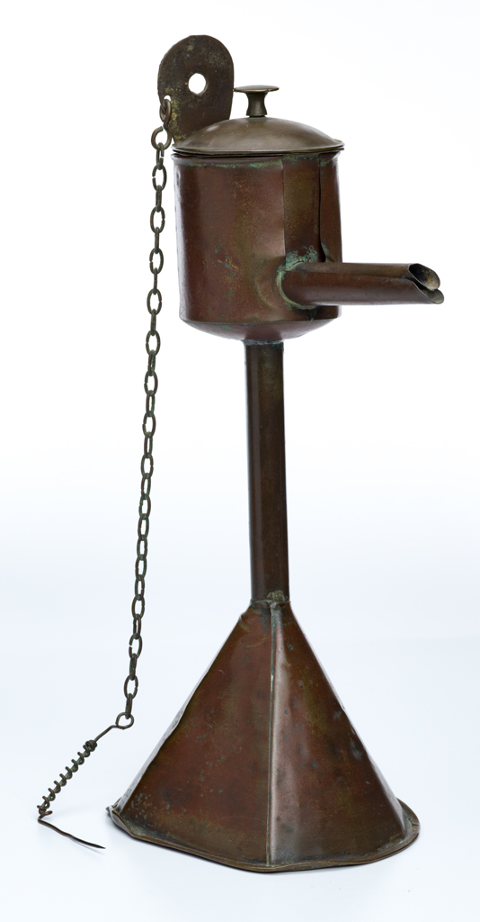 An oil lamp made of metal, has a seperate round central oil container with lid and a spout at one end where the tip of an oil soaked wick could protrude.  A pick, for rescuing the wick is on the end of a chain attached at the top of the main base. The base is a funnel with one flattened side.