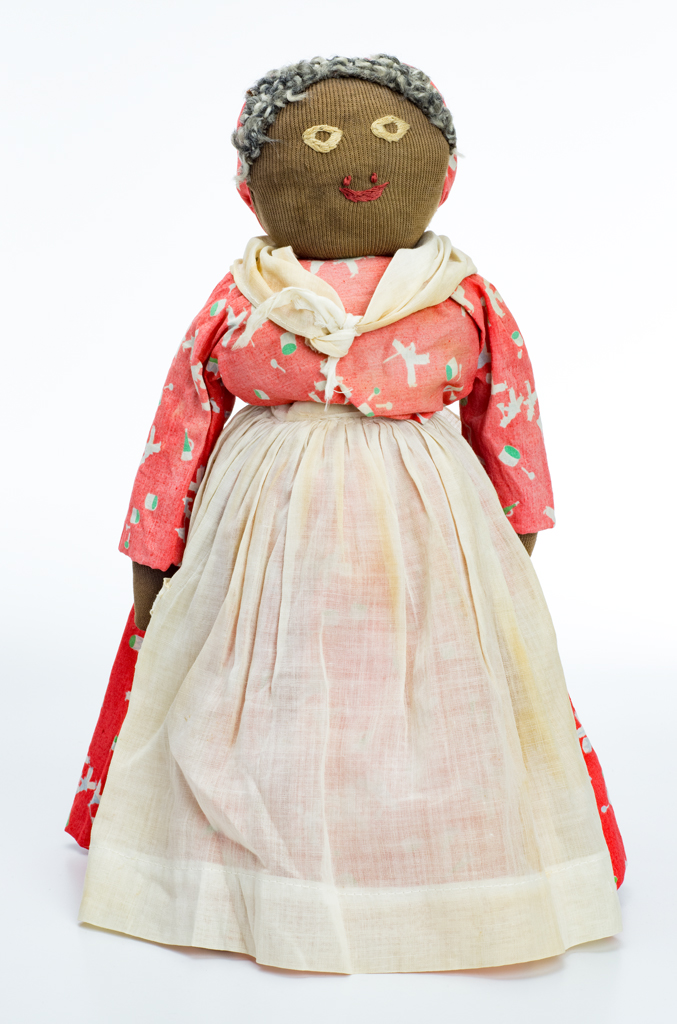 A female bottle doll with embroidered eyes and mouth. She has a pink printed dress, covered by a white cotton apron and scarf. Her yarn hair is covered with a pink hankerchief.