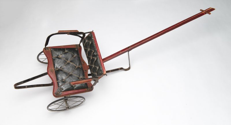 Metal and wood cart with a long handle, two wheels, and a small seat. The wood elements are painted red with gold decoration on the seat back and the metal elements are painted black. The seat has two padded fabric cushions tacked into place with brass tacks. The fabric has a black coating to resemble leather. The metal wheels are fitted with solid rubber strips to create tires.