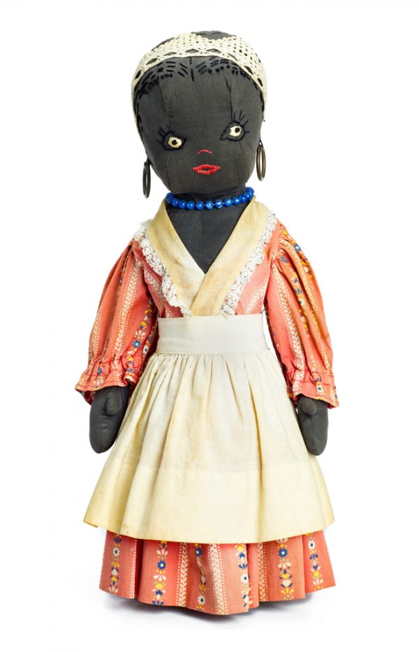 A female bottle doll wears a pink printed dress, large hoop earrings and a blue bead necklace. Her apron is white cotton with cotton crocheted lace edging. Her head covering is a wide band of crocheted lace. Her features are embroidered.