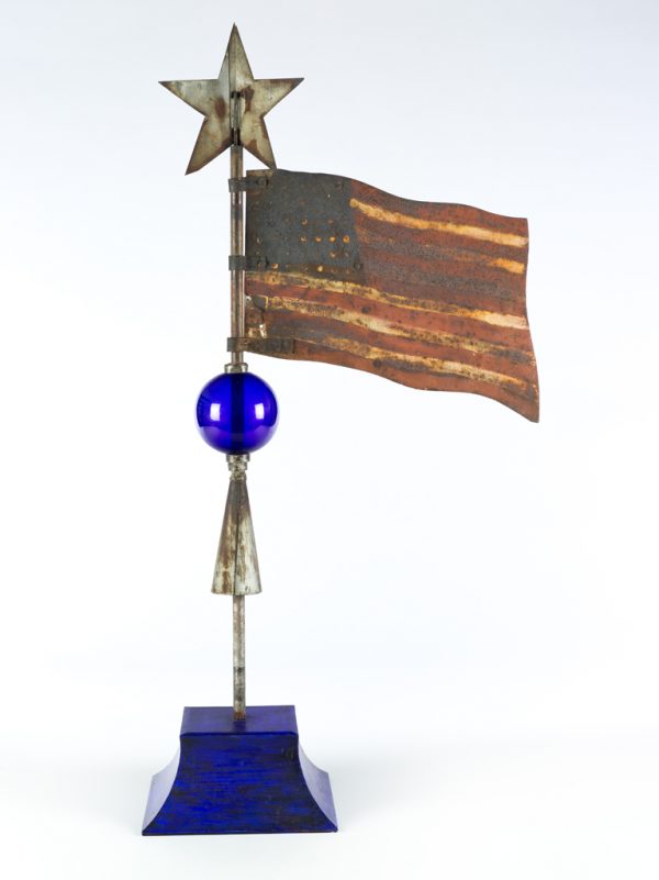 An American flag weathervane. The flag is mounted to a post. A blue sphere of glass is blown and mounted to the flag post just below the flag, and a three-dimensional star is atop the post.