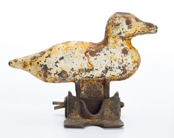 cast iron duck in relief, which is mounted to a cast iron bracket. The duck is painted white and paint traces are present on the bracket. The duck pivots on the bracket and would tip over when hit as part of a carnival game.