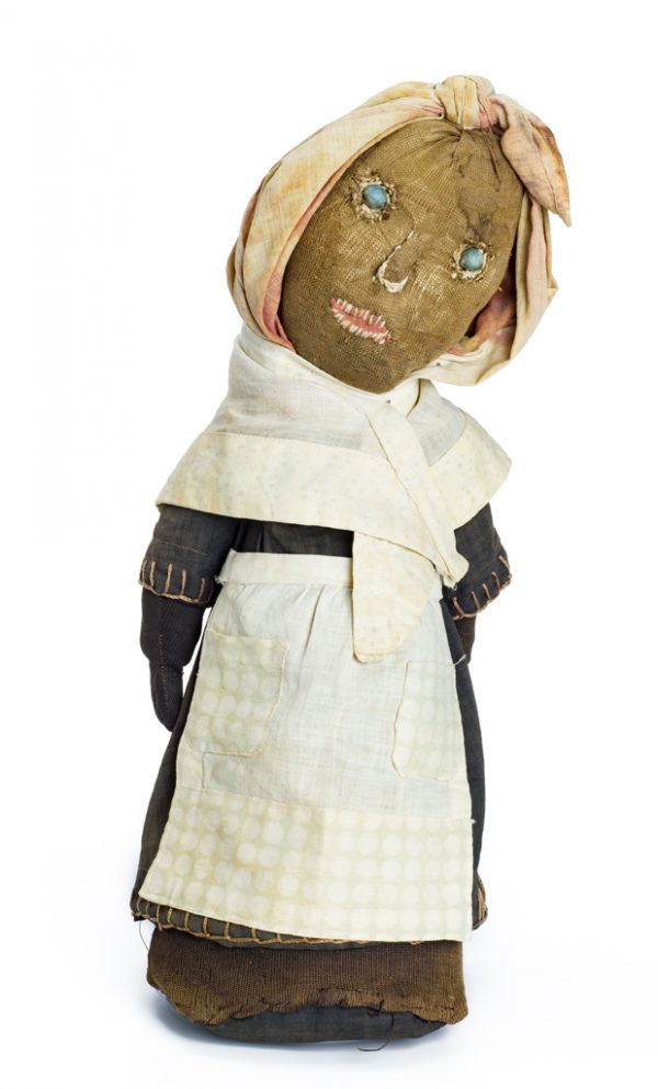 A female black doll with cloth head scarf. Her features are embroidered with solid eyes (cloth over bead). Her brown dress is covered with a white cotton (printed and checkered) shawl and apron. Her body functions like a bottle filled with sand, but the straw fill won't support the doll. Made by same maker as 2016.1.11