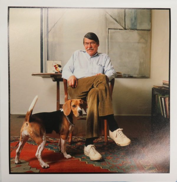 The artist Richard Diebenkorn sits in front of a painting.  A dog faces him, but looks toward the viewer.