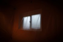 A plain orange colored room, has at the center a window with blurred curtains blowing in the wind..
