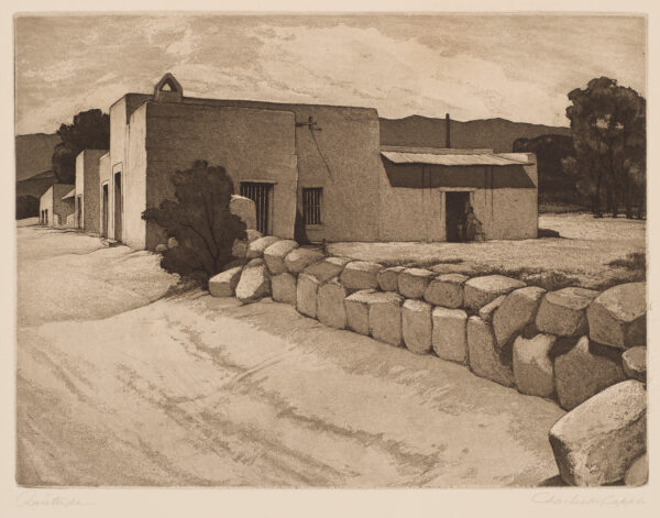 A woman stands with a chair in the doorwar of an adobe building with bars on the windows. Large rocks form a fence along the road to the left.