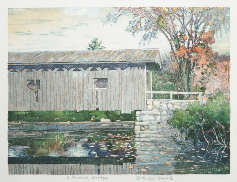 A covered bridge stands over a reflective lake with fall colored trees in the distance.