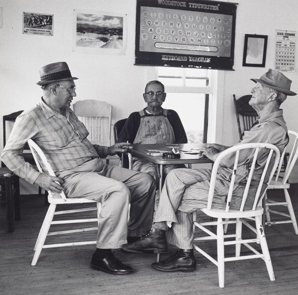 Three older men sit at square table with three white painted chairs and a black chair playing cards. Two men wear hats, and one is smoking a cigarette. The background has posters, most prominent is a key board diagram. Window and additional chairs behind the men.