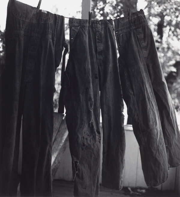 Three pairs of overalls hanging by the waist from a clothesline, held up with clothes pins with windows in the background