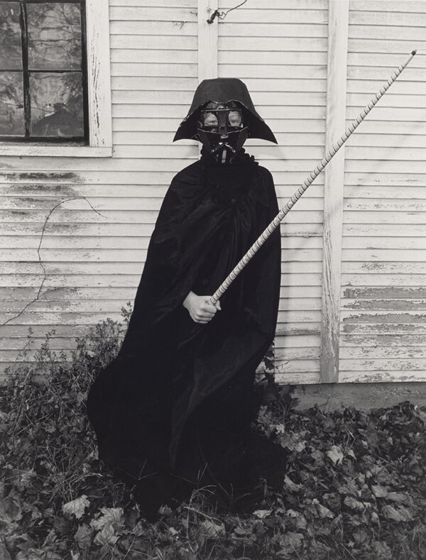 Young boy (Sage Schwarm) dressed up in homemade Darth Vader costume with light saber looking at viewers as he poses against house.