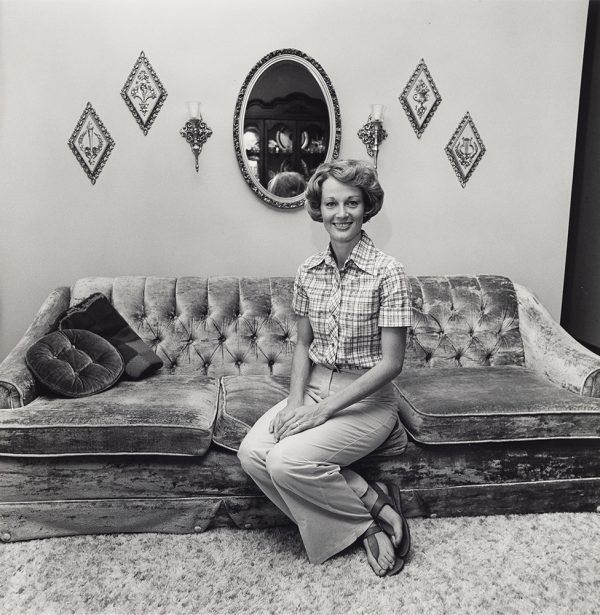 Young woman sits, smiling, with her ankles crossed on a couch with a mirror reflecting a china cabinet, gauntlets, and art pieces behind her on the wall behind her.
