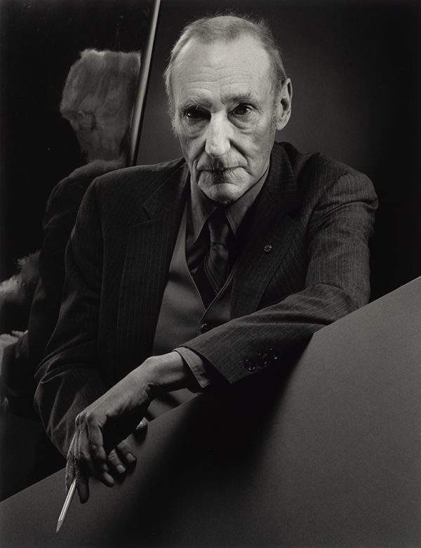 William Burroughs as an older man rests his proper left arm on a flate object, with pencil in his hand. Mirror in the background to the left of figure.