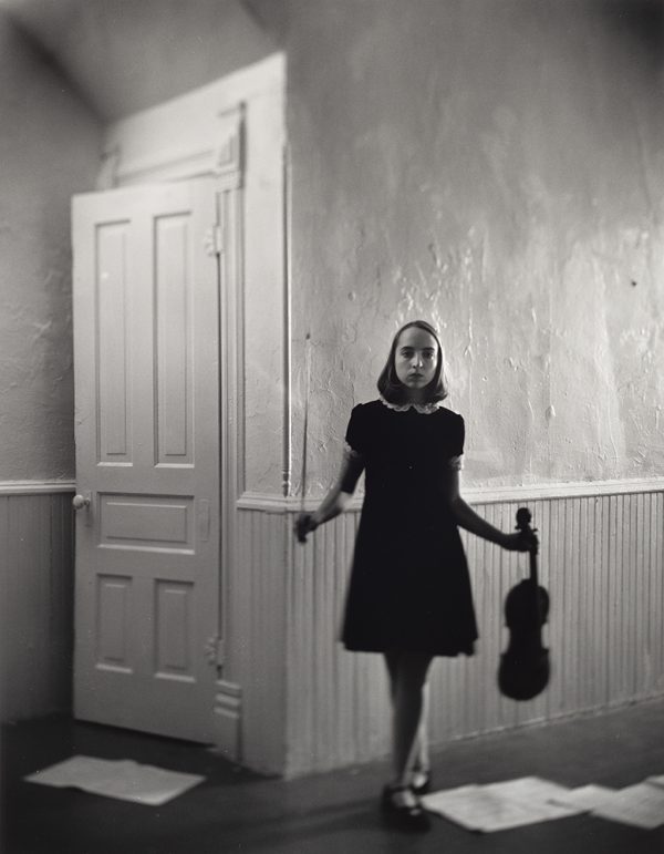 Girl in a black dress stands with a violin in her proper left hand against the background of a light colored room with a slightly open door to the left.