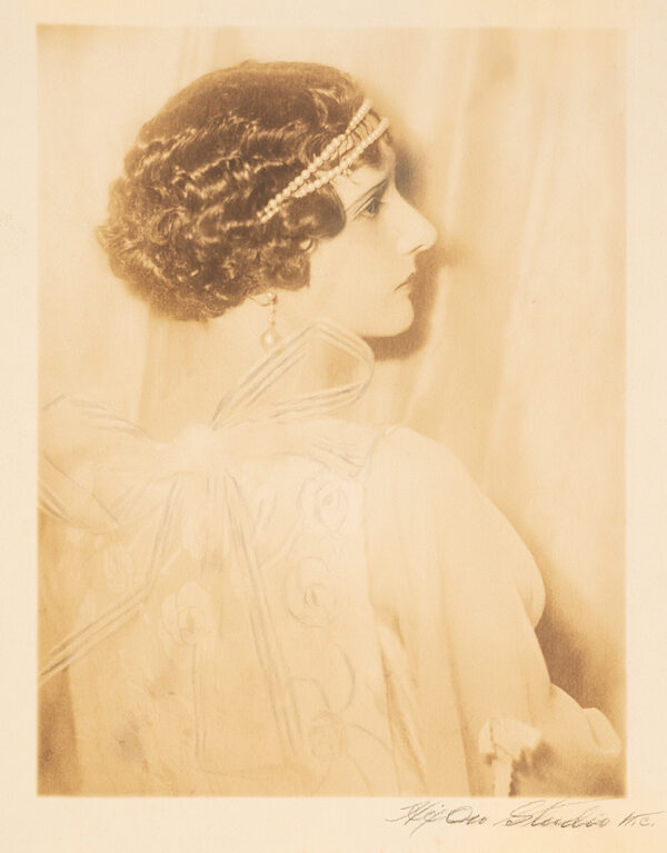 Woman with back to viewers turns her head to the right over her shoulder in profile. She has a large bow on her back, a pearl dangle earring and a headband.
