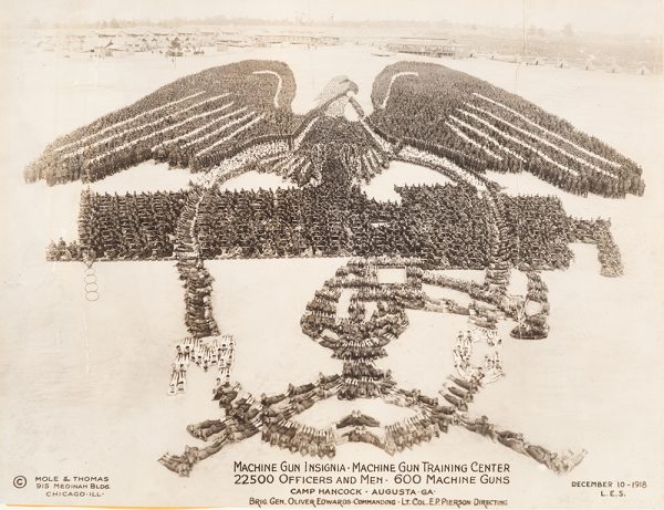 Many officers and men align to create a machine gun insignia when photographed from above.