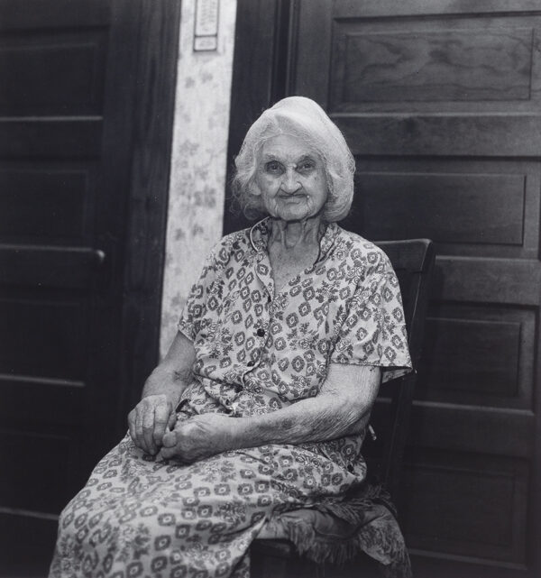 Elderly woman sitting in wooden chair with 2 doors behind her.