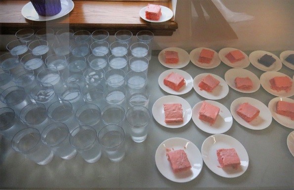 Multiple rows of glasses half-full of water, and plates of frosted pink cake.