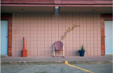 An exterior pink motel wall, with single lawn chair looking toward the parking lot. A door is on each side.