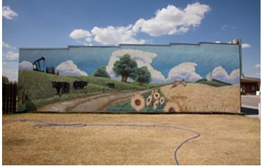A mural of an oil derrick on the left, cows, a road and wheat field with sunflowers at bottom center is seen against a blue sky with white clouds.