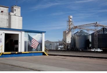 Flag mural on Co-Op Exchange building, cement lot, industrial farming equipment in the background.